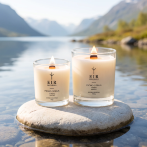Velas aromáticas Fjord Citrus da EIR Naturals, 50g e 190g acesas sobre pedra clara à beira da água cristalina de um fiorde