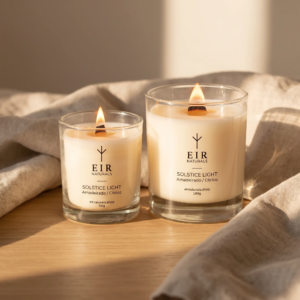 Velas aromáticas Solstice Light da EIR Naturals, 50g e 190g acesas sobre madeira clara com linho natural em luz de fim de tarde